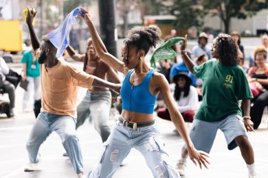A group of dancers holding colourful scarves perform outdoors in front of a live audience.