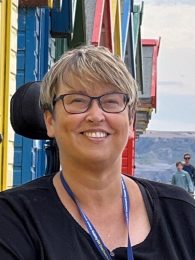 Headshot of person with short blonde hair, wearing a black shirt with a blue lanyard, smiles at the camera.