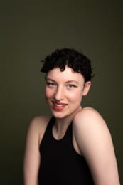 Headshot of a person with short, curly hair, warmly smiling at the camera against a dark green background.