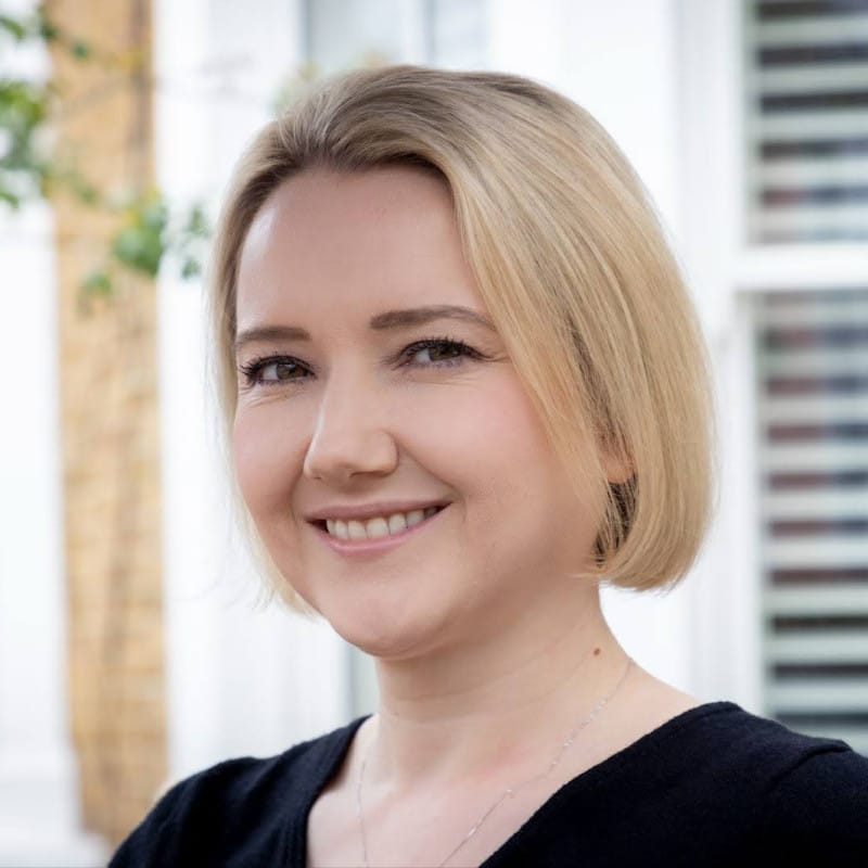A warm, natural headshot of a person with straight blonde hair styled in a sleek bob cut, smiling gently at the camera. The photo is taken outdoors in soft, natural light, with a blurred background.