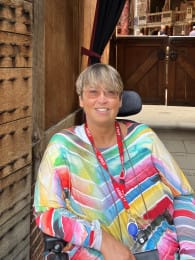 Sarah Howard, smiling at the camera, wearing light-tinted sunglasses and a bright, multicoloured striped dress in pink, green, yellow, and blue. Sarah has short blond hair and a red lanyard around their neck.