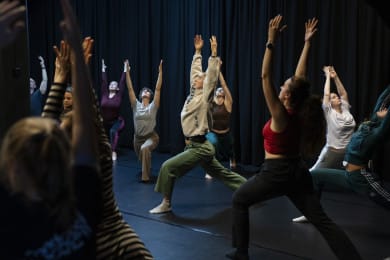 A group of people practising stretching or yoga in a dimly lit indoor space. They are leaning forward with one leg extended and arms reaching upward.