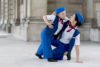 Two performers in matching blue uniforms pose mid-movement in a bright outdoor setting, leaning into each other with focused, connected expressions.
