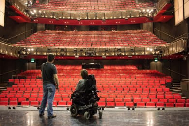 Two people are on an empty stage with their backs to us, looking out into an auditorium filled with red velvet seats. The person on the left wears jeans and has a casual stance. The person on the right is seated in an electric wheelchair.