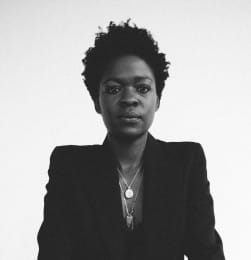 A black-and-white portrait of a person with short textured hair, wearing a dark jacket and layered necklaces, looking directly at the camera.