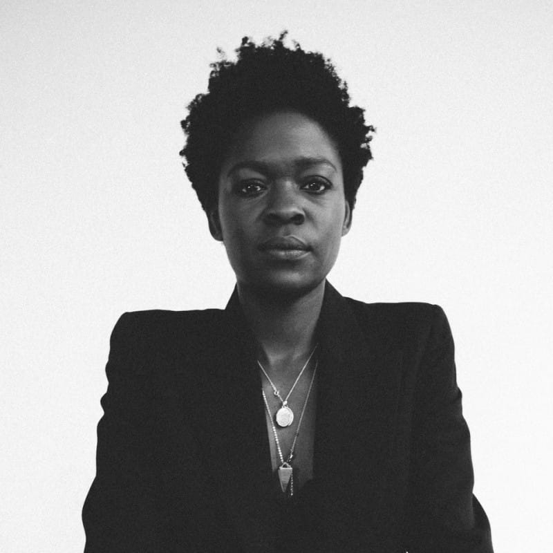 A black-and-white portrait of a person with short textured hair, wearing a dark jacket and layered necklaces, looking directly at the camera.