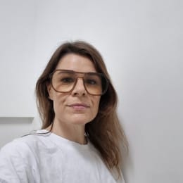 A person with long brown hair wears large glasses and a white top, looking at the camera against a plain background.