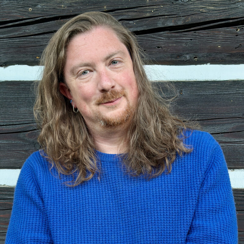 A person with long light brown hair and a neatly trimmed moustache and goatee smiles gently at the camera while wearing a bright blue textured sweater. They are standing in front of a weathered dark wooden wall with white horizontal trim, giving the portrait a warm, relaxed feel.