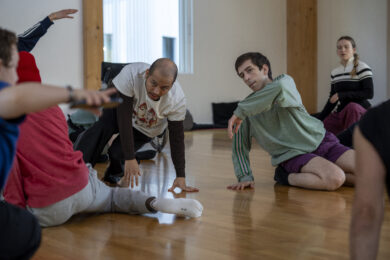 Several people are gathered in a dance studio, seated or crouched on the wooden floor as they take part in a movement session. Two participants in the centre are captured in expressive, low-to-the-ground poses, while others watch, respond or prepare to move, creating a sense of shared attention and collaboration. The bright, open space with timber columns and natural light reinforces the atmosphere of exploration and collective learning.