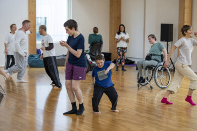 A group of people are moving and dancing together in a bright studio with a wooden floor, creating an atmosphere of shared exploration and creativity. Some participants are standing, others are using wheelchairs or seated mobility devices, and everyone is engaged in their own movement while sharing the space. Large windows and timber columns fill the room with natural light, emphasising the open, welcoming environment.